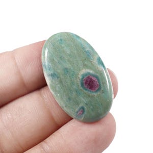 Könnte beinhalten: Oval geformter Edelstein mit polierter Oberfläche. Der Stein ist hauptsächlich in einem gedämpften Grün gehalten, mit dunkleren grünen und rot-violetten Einschlüssen. Der Edelstein wird in der Hand einer Person gehalten.