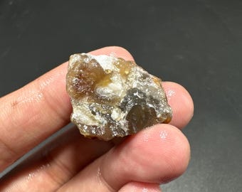 May include: A rough, uncut gemstone with a mix of brown, tan, and white colors. The stone has a natural, irregular shape and a slightly translucent appearance. The image shows the stone held in a hand, highlighting its texture and color variations.