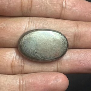 Könnte beinhalten: Ein silberfarbener, oval geformter Stein mit glatter, polierter Oberfläche. Der Stein hat eine leicht unregelmäßige Textur. Der Stein ist wahrscheinlich ein Edelstein oder ein dekoratives Element. Der Hintergrund ist neutral.