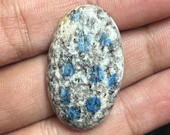 May include: Oval-shaped K2 stone cabochon. The stone features a speckled pattern of white, gray, and blue. The blue spots are scattered throughout the stone, creating a unique and natural design. This polished stone is ideal for jewelry making.