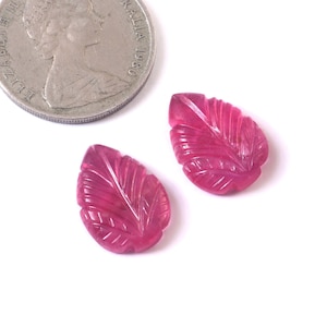 Peut inclure: Deux pendentifs en forme de feuilles en pierres précieuses de couleur rubis sculptées. Les pierres précieuses sont à côté d'une pièce de monnaie en argent. Les feuilles ont des veines détaillées et sont posées sur un fond blanc. Les pierres précieuses mesurent environ 2,5 cm de long.