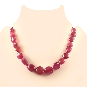 Puede incluir: Un collar con piedras preciosas pulidas, de forma ovalada y color rubí. Las piedras varían en tamaño y tono, desde el rojo intenso hasta los tonos rosados más claros. El collar se exhibe en un maniquí.