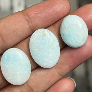 May include: Three oval-shaped gemstones with a pale blue and white color scheme are displayed. The stones have a smooth, polished surface and a marbled appearance. The gemstones are held in a hand, showcasing their size and color.