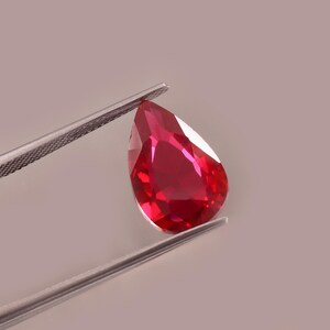 Op de afbeelding: Een peervormige, gefacetteerde robijn, vastgehouden door een zilveren pincet. De dieprode edelsteen toont interne reflecties, wat de helderheid benadrukt. De achtergrond is een zachte, neutrale tint.