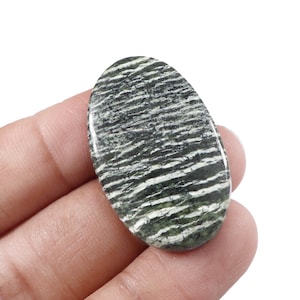 Könnte beinhalten: Ovaler Edelstein mit einem gestreiften Muster in Grün und Weiß. Der polierte Stein wird in einer Hand gehalten, wodurch seine glatte Oberfläche und natürlichen Variationen hervorgehoben werden. Das Design des Edelsteins ähnelt einer Landschaft.