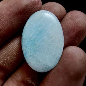 May include: Oval-shaped, polished gemstone with a light blue hue and subtle white veining. The stone's surface is smooth and reflective, with a soft, cloudy appearance. The gemstone is held in a hand, showcasing its natural beauty and unique patterns.