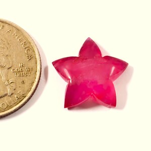 Peut inclure: Une pierre précieuse en forme d'étoile aux tons roses et magenta. La surface polie reflète la lumière, mettant en valeur la qualité translucide de la pierre. Une pièce de monnaie américaine est à côté pour la comparaison.