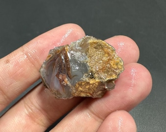 May include: A rough, uncut gemstone with a mix of colors, including brown, tan, gray, and white. The stone has a natural, organic shape and texture, with visible layers and variations in color. The gemstone is held in a hand.