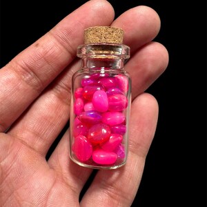 May include: A small, clear glass bottle filled with vibrant pink and magenta gemstones. The bottle has a cork stopper and is held in a hand against a black background. The gemstones appear smooth and polished.