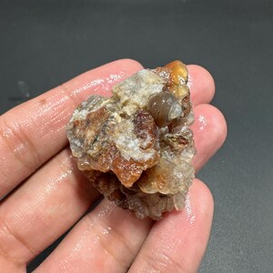 Puede incluir: Una gema en bruto, sin cortar, con una mezcla de colores, incluyendo tonos marrones, naranjas y blancos. La piedra en bruto tiene una superficie texturizada y una forma irregular, mostrando su estado natural, sin pulir. La gema se sostiene en una mano.