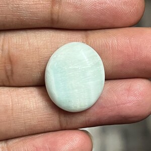 May include: A pale blue, oval-shaped gemstone with a smooth, polished surface. The stone has subtle, light-colored veining throughout. The gemstone is held in a person's hand, showcasing its natural color and texture. This gemstone is likely used for jewelry.