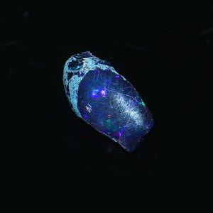 May include: A polished, elongated opal gemstone with a dark blue base color. The opal has iridescent flashes of color, including blue, green, and red. The top edge has a light blue, rough texture. The gemstone is set against a black background.