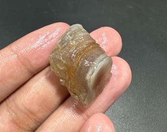 May include: A raw, natural stone with a layered, banded appearance. The stone features a mix of tan, brown, and white colors, with a slightly translucent quality. The texture appears rough and organic, showcasing natural geological formations.