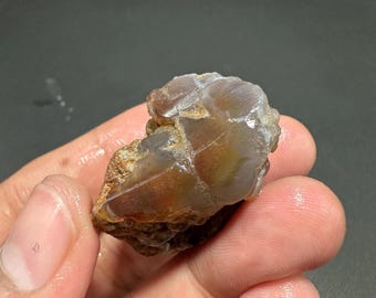 May include: A raw, uncut gemstone with a mix of brown, tan, and translucent areas. The stone has a rough, natural texture and is held in a hand, showcasing its organic form. The gemstone appears to be a type of agate or similar mineral.