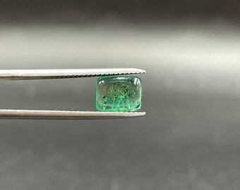 May include: A green emerald gemstone, cut into a square shape, is held in tweezers. The gemstone is transparent and has a slight sparkle.