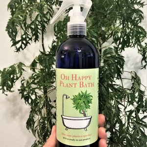 May include: A blue bottle with a clear spray nozzle, labeled "OH HAPPY PLANT BATH". The label features a cartoon illustration of a plant in a bathtub. The text on the label reads "Give your plants a spa day! 16 fl oz ready-to-use product."