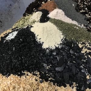 May include: A close-up of a mixture of different soil amendments, including black charcoal, brown compost, white perlite, and green vermicast. The soil amendments are arranged in a pile, with the charcoal and compost being the most prominent.