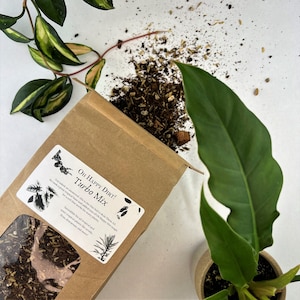 May include: A brown paper bag with a label that reads "OH HAPPY DIRT! Turbo Mix" and a description of the product. The bag is filled with a dark brown soil mix. A green plant is in the background.