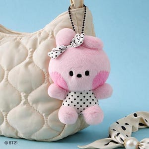 May include: A plush pink character keychain with black eyes and a nose, wearing a white and black polka dot outfit and a matching bow on its head. The soft toy is attached to a black ball chain and features the text BT21 printed in the corner.