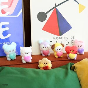 Könnte beinhalten: Eine Sammlung von sieben Plüschfiguren in verschiedenen Pastellfarben, jede mit einem Herz. Die Spielzeuge haben verschiedene tierähnliche Designs, darunter Bären und Hasen. Der Hintergrund umfasst eine grüne Oberfläche und ein Poster mit abstrakter Kunst.