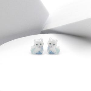 May include: A pair of white cat stud earrings with blue and green watercolor details. The cat is sitting with its head tilted slightly to the side.