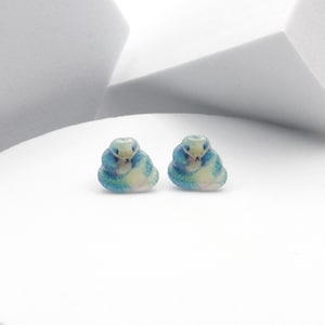 May include: A pair of small, snake-shaped earrings. Each earring is a coiled snake with a blue and white colour scheme. The earrings are set against a white background.
