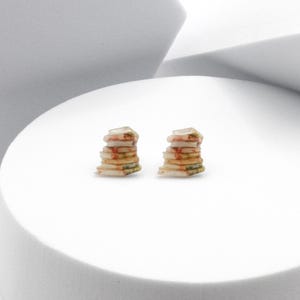 Peut inclure: Une paire de petites boucles d'oreilles en forme de livre. Chaque boucle d'oreille est une pile de cinq livres miniatures aux couvertures colorées. Les livres sont en résine et ont un aspect réaliste.
