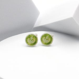 May include: Two round silver cufflinks featuring a green and white latte art design. The cufflinks are presented on a white surface, set against a backdrop of geometric shapes.