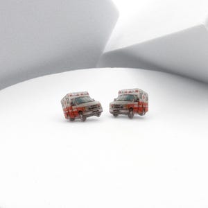 May include: A pair of miniature ambulance earrings. Each earring features a detailed replica of an ambulance, with a white body, red accents, and emergency lights. The earrings are set against a white background.