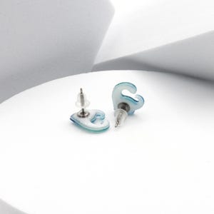 Puede incluir: Un par de pendientes azules y blancos con forma de ola. Los pendientes est&aacute;n hechos de un material transl&uacute;cido y tienen un poste plateado.
