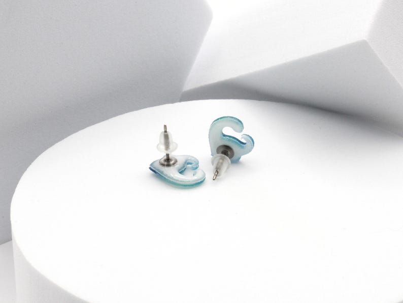 Puede incluir: Un par de pendientes azules y blancos con forma de ola. Los pendientes est&aacute;n hechos de un material transl&uacute;cido y tienen un poste plateado.