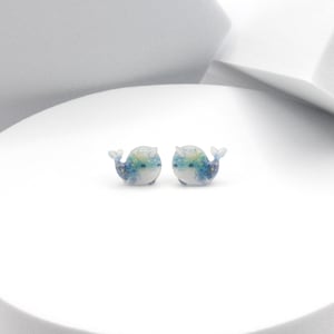 Può includere: Un paio di orecchini a forma di balena, piccoli e stravaganti. Ogni orecchino presenta una base bianca con dettagli blu, verdi e gialli e una piccola coda. Gli orecchini sono esposti su una superficie bianca e arrotondata.