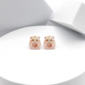 Könnte beinhalten: Ein Paar kleine, rosafarbene Hamster-Ohrringe. Jeder Ohrring zeigt einen Hamster, der ein kleines, braunes Objekt in seinen Pfoten hält.