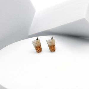 Op de afbeelding: Een paar oorbellen in de vorm van bubble tea bekers. De oorbellen tonen een lichtbruine beker met een witte bovenkant, een rietje en bruine tapiocaparels. Op een witte achtergrond.
