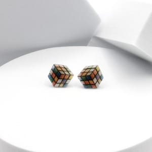 Puede incluir: Un par de pendientes de madera coloridos con forma de cubo de Rubik. Cada pendiente es un pequeño cubo multicolor con líneas negras que delimitan cada cuadrado.
