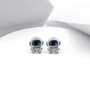 Puede incluir: Un par de pendientes de botón de robot blanco con detalles azules. Los robots tienen ojos negros y miran hacia adelante.
