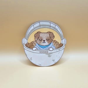 May include: A cartoon illustration of a dog in a baby carrier. The dog has brown fur and is wearing a blue shirt. The carrier is white with a gray handle and a light brown background.