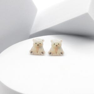 May include: Two small, cream-colored bear figurines with brown details, sitting side-by-side on a white surface. The bears have small, dark eyes and are posed in a seated position. The background is a minimalist white geometric design.