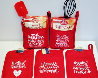 Op de afbeelding: Vier rode ovenwanten met witte tekst. De wanten hebben de volgende tekst: "baked with love by MOM", "Happiness is homemade", "Christmas Calories Don't Count" en "Thanks for making me one smart cookie". Twee rode stoffen tassen met witte tekst. De tassen hebben de volgende tekst: "We whisk you a Merry Christmas" en "Christmas Calories Don't Count". De tassen bevatten dozen Betty Crocker suikerkoekjesmix en Betty Crocker walnoot-chocoladekoekjesmix. Een rode spatel en een zwarte garde zijn ook inbegrepen.