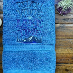 May include: A blue bath towel with the embroidered text "Relax You're Lake Time" and a mountain scene with trees.