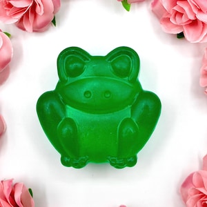 May include: A translucent green frog-shaped soap bar. The soap has a detailed frog design with visible eyes, legs, and feet. The soap is surrounded by pink roses on a white background.