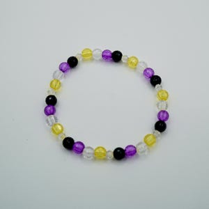 May include: A beaded bracelet featuring a non-binary pride flag color scheme. The bracelet is composed of small, round beads in clear, yellow, purple, and black. The beads are strung together to form a circular shape.
