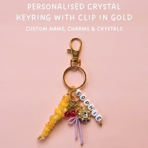 May include: Gold-colored keyring with a clip, featuring a cluster of yellow crystals, red beads, and a charm with the name "SOPHIE" spelled out in white beads. The text on the image reads "PERSONALISED CRYSTAL KEYRING WITH CLIP IN GOLD".