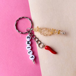 May include: A silver keyring with the name "EMILY" spelled out in white letter beads. It features a red chilli pepper charm and a cluster of yellow gemstone chips. The background is a mix of pink and beige.