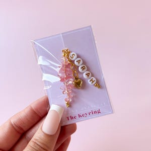 May include: A pink and gold keychain with the name "SARAH" spelled out in white beads. The keychain also features a small gold heart charm and a pink crystal charm.