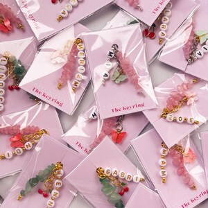 May include: A collection of personalized keychains with different names, each featuring a charm with a butterfly and a cherry. The keychains are made with pink and green beads and are displayed on pink cards with the text "The Keyring".