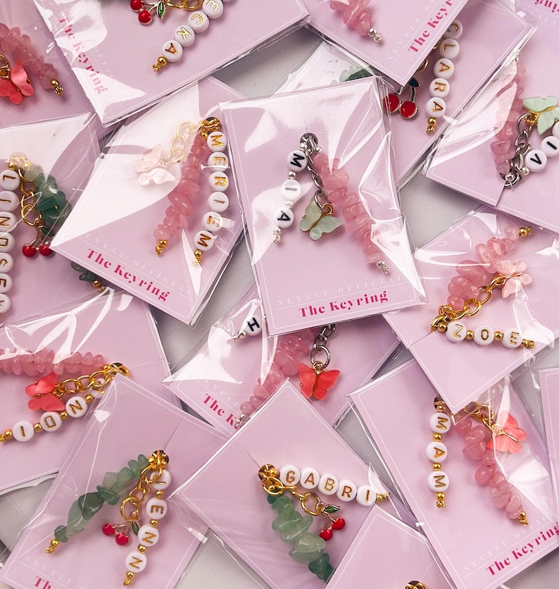 May include: A collection of personalized keychains with different names, each featuring a charm with a butterfly and a cherry. The keychains are made with pink and green beads and are displayed on pink cards with the text "The Keyring".