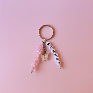 May include: A pink and white keychain with a silver key ring, a pink crystal charm, a white butterfly charm, and a white bead charm with the word "SUMMER" spelled out.
