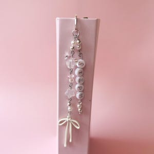 Op de afbeelding: Een witte en zilveren hanger met de naam "Stephanie" gespeld in witte kralen. De hanger heeft ook een witte strik, een witte ster en heldere en roze kralen.