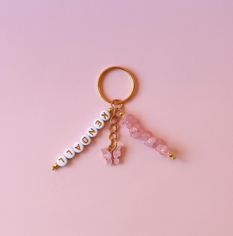 May include: Gold keychain with a pink butterfly charm, a pink crystal charm, and a white beaded charm with the name "KENDALL" spelled out.